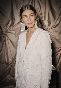 Balta, tekstūruota striukė su užuolaidiniais deimantais, turinti fringing detales ir vieną mygtuko užsegimą, dėvima ant nėrinių viršaus.