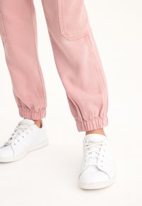 Svetle růžové joggers s elastickými manžetami vyrobené z měkké tkaniny; nošené s bílými teniskami s jednoduchým designem a texturovanou gumovou podrážkou.