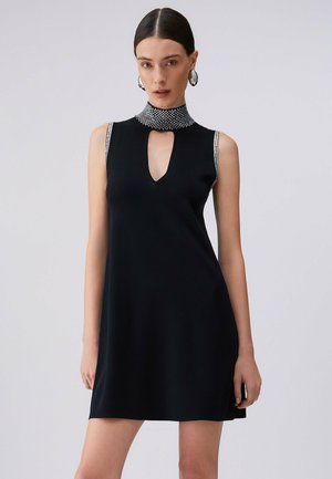 Donna che indossa un vestito nero senza maniche con bordatura di strass sul colletto e sugli spacchi delle braccia, in piedi davanti a uno sfondo chiaro uniforme.