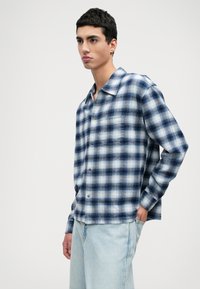 Μπλε και λευκό καρό πουκάμισο με χαλαρή γραμμή, κουμπιά μπροστά και τσέπη στο στήθος. Φτιαγμένο από μαλακό ύφασμα με λεία υφή.