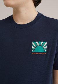 Tums zila kokvilnas T-kreklā ar priekšējā krūtīs grafiku, kas attēlo sauli ar stariem tirkīza un baltā krāsā, ar uzrakstu "BLUE RIDGE DENIM" oranžā krāsā.