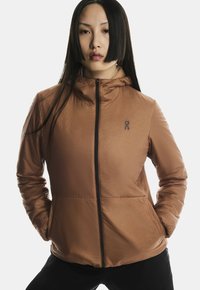 Femeie cu păr drept negru, purtând o jachetă maro cu fermoar, având mâinile în buzunare, stând în fața unui fundal deschis, simplu.