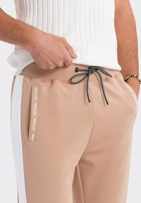 Beige sportieve joggingbroek met een trekkoord in de taille, zijzakken en witte zijstrepen. Heeft een gestructureerde stof en logo-accenten.