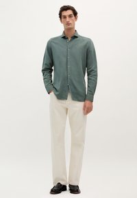 Camisa de lino verde de botones con un corte slim, combinada con pantalones color crema y mocasines negros, contra un fondo claro y liso.