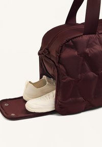 Burgundi, steppelt utazótáska cipzáras rekesszel, melyben fehér sportcipők láthatók. Sima szövet textúra, erős hordozófülekkel.