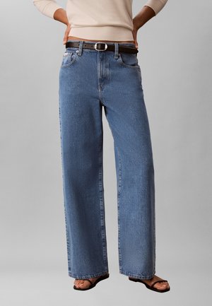 Persona che indossa jeans blu a vita alta e gamba larga con cintura nera, top beige a maniche lunghe e sandali neri, in piedi con le mani sui fianchi.