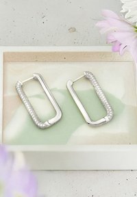 Silver rektangulära örhängen med en texturerad yta och kristallaccenter, visade på en grön och krämfärgad bricka, med blommönster.