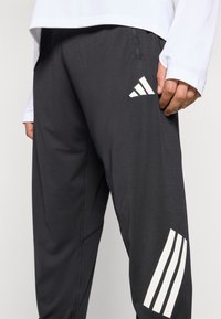 Pantalon de sport noir à texture lisse, avec logos blancs Adidas et trois bandes sur les côtés. Matière extensible et design minimaliste.