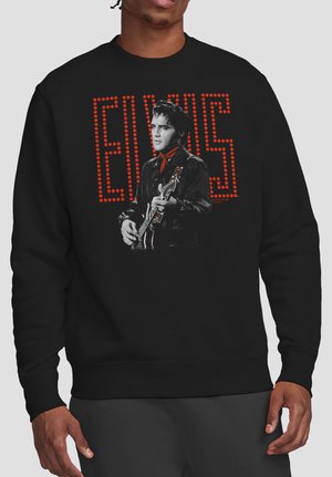 Man wearing black sweatshirt with black-and-white photo of Elvis playing guitar and red dotted text spelling "ELVIS" behind him.