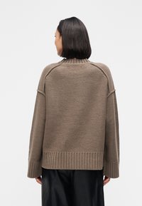 Υπερμεγέθη καφέ πουλόβερ με ριμπ τελείωμα και μανσέτες, πλεκτό σχέδιο με υφή και ψηλό λαιμόκοψη. Η πίσω όψη έχει ομοιόμορφο φινίρισμα χωρίς ραφές και χαμηλωμένους ώμους.