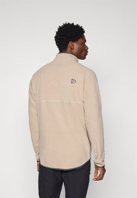 Pullover in pile beige con colletto alto, dettagli grigi a contrasto sui polsini e un piccolo logo sulla parte posteriore. Texture liscia, vestibilità rilassata.
