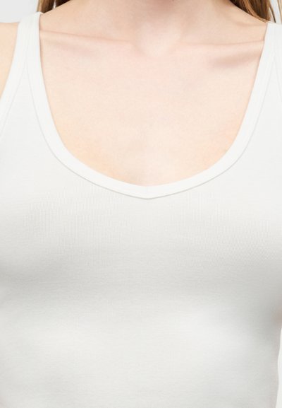 Κοντινό πλάνο γυναίκας που φοράει λευκό ριμπ αμάνικο τοπ με στρογγυλή λαιμόκοψη, που δείχνει τον λαιμό και το πάνω μέρος του στήθους της.