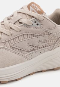 Beige sportcipő velúr és hálós felsőrésszel, texturált kiegészítőkkel, lapos fűzőkkel és gumitalppal. A nyelvén márkás címke található.