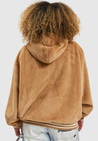 Beżowa pluszowa kurtka z kapturem o miękkiej fakturze, obniżonych ramionach i ściągaczu na dole. Posiada przytulny kaptur i luźny fason.