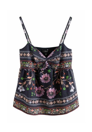 Jeune femme aux cheveux ondulés portant une robe noire sans manches à motif floral, appuyée contre un mur uni de couleur claire.