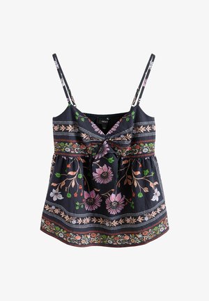 Jeune femme aux cheveux ondulés portant une robe noire sans manches à motif floral, appuyée contre un mur uni de couleur claire.