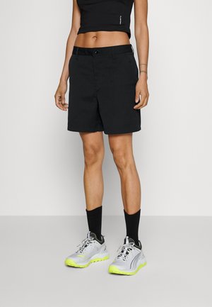 Person iført sorte knælange shorts, sort crop top, sorte sokker og lysegrå sneakers med gule såler, står mod en ensfarvet baggrund.
