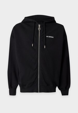 Jonge man staand, draag een zwarte hoodie met rits, zwart T-shirt met witte tekst, losse blauwe spijkerbroek en zwarte sneakers tegen een effen achtergrond.
