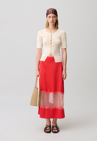 Donna che indossa una maglietta crema a maniche corte con bottoni, gonna rossa con pannello di pizzo, sandali marroni, foulard marrone a pois e tiene in mano una borsa tote beige.