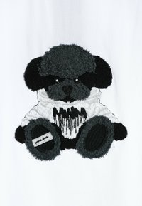 Osito de peluche en negro y gris, con pelaje texturizado, una camiseta blanca con detalles en negro, y orejas y pies redondeados y distintivos.