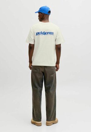 Homme portant un T-shirt blanc avec le logo bleu "jack & jones" dans le dos, pantalon marron, bottines beiges et casquette de baseball bleue, debout de dos.
