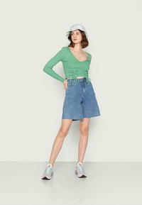 Top de manga longa verde com pregas laterais e cordões, combinado com calções de ganga azul médio. A modelo usa ténis desportivos coloridos e um chapéu tipo balde.