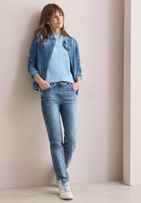 Džínová bunda v světle modré barvě s žebrovanými manžetami, v kombinaci se svetrem v světle modré barvě a středně modrými džínami. Bílá sneakers dokončují vzhled.