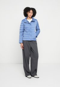 Hele sinine, tepitud muhviga jakk, millel on püstitatud krae, nööbid ja küljetaskud, sobitatud hallide laia lõikega pükstega ning valgete kingadega.