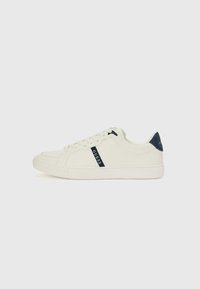 White canvas sneaker with navy logo detail, round toe, lace-up design, and flat rubber sole. Features a suede heel accent in navy.