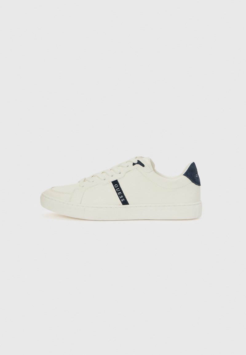 White canvas sneaker with navy logo detail, round toe, lace-up design, and flat rubber sole. Features a suede heel accent in navy.