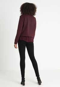 Veste en maille bourgogne avec tissu texturé, ourlet arrondi et manches longues ; associée à un pantalon noir ajusté et des bottines à talons.