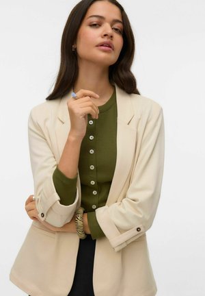 Blazer beige porté sur un haut côtelé vert avec des détails de boutons, manches retroussées, accessoirisé d'un bracelet à motifs.
