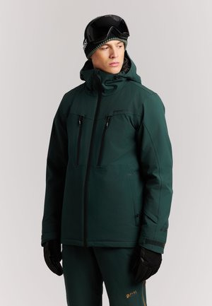 Man draagt een donkergroene skipak en broek met zwarte handschoenen, een grijze muts en een zwarte skibril, en kijkt opzij tegen een lichte achtergrond.