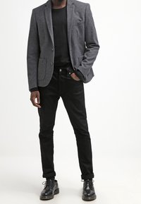 Šedý vlněný blazer s dvěma předními kapsami, nošený přes černou košili. Černé slim-fit džíny a černé šněrovací boty dotvářejí outfit.