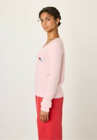 Pull en peluche rose clair avec un décolleté en V profond, orné d'un petit patch coloré sur la poitrine gauche. Assorti à une jupe rouge.