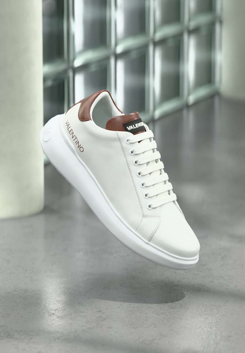 Λευκό δερμάτινο αθλητικό παπούτσι με καφέ δερμάτινες λεπτομέρειες, στρογγυλή μύτη, επίπεδη λευκή σόλα, έξι τρύπες για κορδόνια και ανάγλυφο λογότυπο στη φτέρνα.