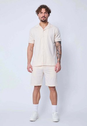 Homme aux cheveux bouclés et tatouages portant une chemise beige clair à manches courtes boutonnée, un short assorti, des chaussettes blanches et des baskets blanches, debout.