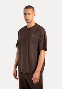 Brun bomulds T-shirt med rund halsudskæring og korte ærmer, med et lille logo mærke foran. Glat tekstur.