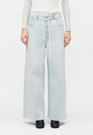 Kvinde iført lyseblå bukser med vide ben og hvid snor, kombineret med en hvid langærmet top og sorte sko.