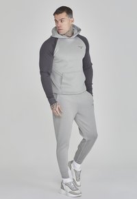 Felpa sportiva grigia con maniche nere, caratterizzata da una tasca frontale e un logo. Abbinata a pantaloni jogger grigi e sneakers grigie.