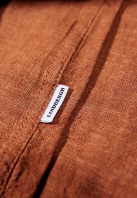 Roestkleurige stof met een gestructureerde weving, met een wit label waarop in zwarte tekst "LINDBERGH" staat. Close-up van het materiaal.