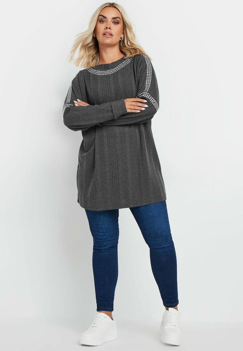 Långärmad grå tunika med texturerad tyg och dekorativa detaljer längs axlarna, ihop med slim-fit mörkblå jeans och vita sneakers.