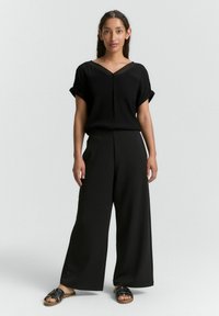 Haut noir à manches courtes avec un col en V et des manches roulées, associé à un pantalon noir à jambes larges. Porté avec des sandales noires à lanières.