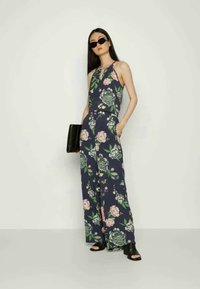 Macacão floral em tecido azul-marinho com grandes flores rosas e verdes, pernas largas e decote halter. Acessórios incluem óculos de sol pretos e uma clutch.