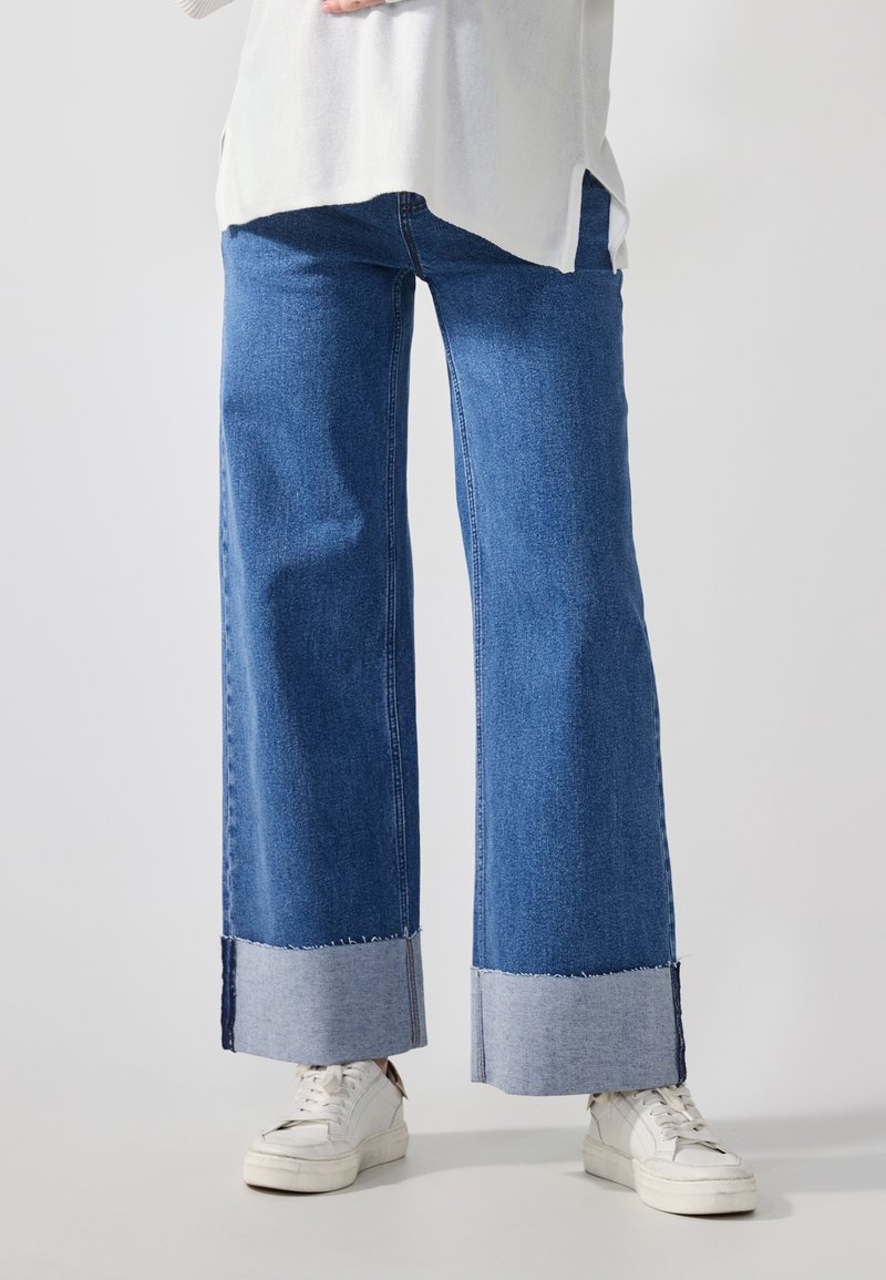 Personne portant un jean bleu à jambes larges avec de grands ourlets retroussés et des baskets blanches, debout devant un fond clair uni.