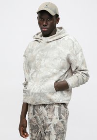 Világosszürke kapucnis pulóver levélmintával, kenguru zsebbel és bordázott mandzsettával, levélmintás nadrággal párosítva, valamint egy bézs sapkával.