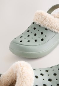 Gröna skumtofflor med en fluffig krämfärgad fuskpälsfoder, med flera ventilationshål och en baksnodd för stöd.