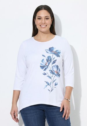 Lächelnde Frau, die ein weißes Langarmshirt mit blauem Blumenmuster vorne und dunkelblaue Jeans trägt, steht vor einem weißen Hintergrund.