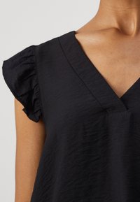 Top nero texture con scollatura a V e maniche corte arricciate. Il materiale sembra leggero e morbido, creando una silhouette rilassata.
