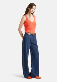 Top corto lavorato a maglia arancione, jeans blu a vita alta con gamba larga e ballerine arancioni a punta. Le caratteristiche visive principali includono texture e colori vivaci.